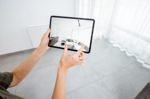 Uma sala branca, com uma cortina branca. Uma pessoa segurando um tablet e na tela do dispositivo a projeção de um sala mobiliada.