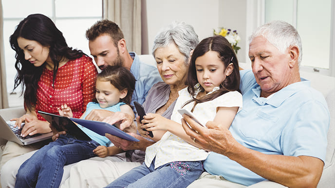 Seis pessoas da mesma família, sendo o casal de avós, os pais e duas crianças, sentados no sofá da sala, usando vários dispositivos móveis, como celulares, tablets e notebook, todos conectados à internet da Desktop.