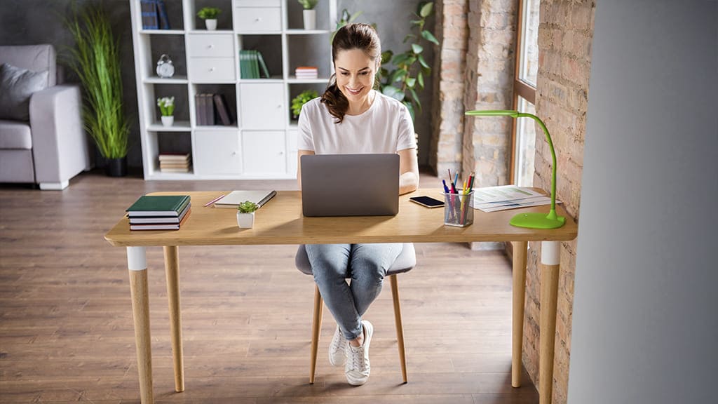 Uma mulher trabalhando na sala de casa. Ela está sentada em uma mesa de escritório de madeira, usando um notebook. Ao seu lado, cadernos, papeis e canetas.