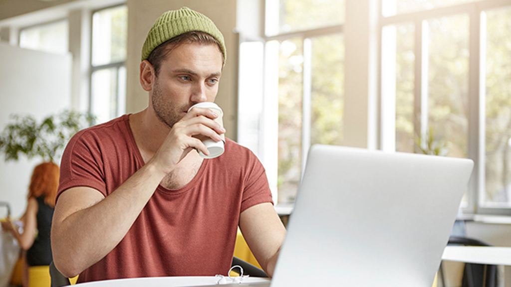 Um homem, em um ambiente interno, sentado à mesa, tomando um café e usando um notebook conectado à internet da Desktop.