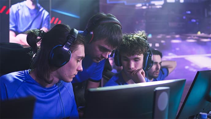 Em um cenário com vários gamers, três meninos estão próximo a dois computadores conversando, dois dos meninos estão sentados e um em pé, curvado.