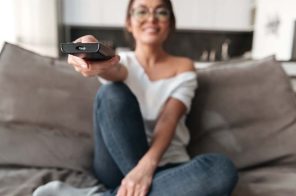 Uma mulher sentada no sofá segurando um controle remoto para ligar a TV.