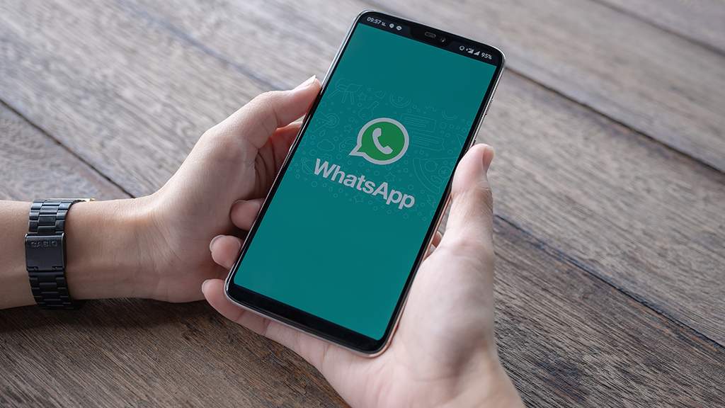 As mãos de uma pessoa posicionada sobre uma mesa de madeira, segurando o celular. Na tela, a imagem principal do app do WhatsApp.