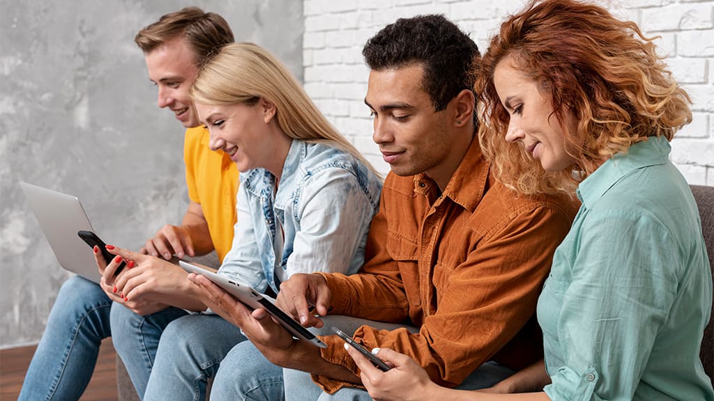 Um grupo de amigos composto por quatro pessoas, todos conectados à internet no notebook, celular e tablet.