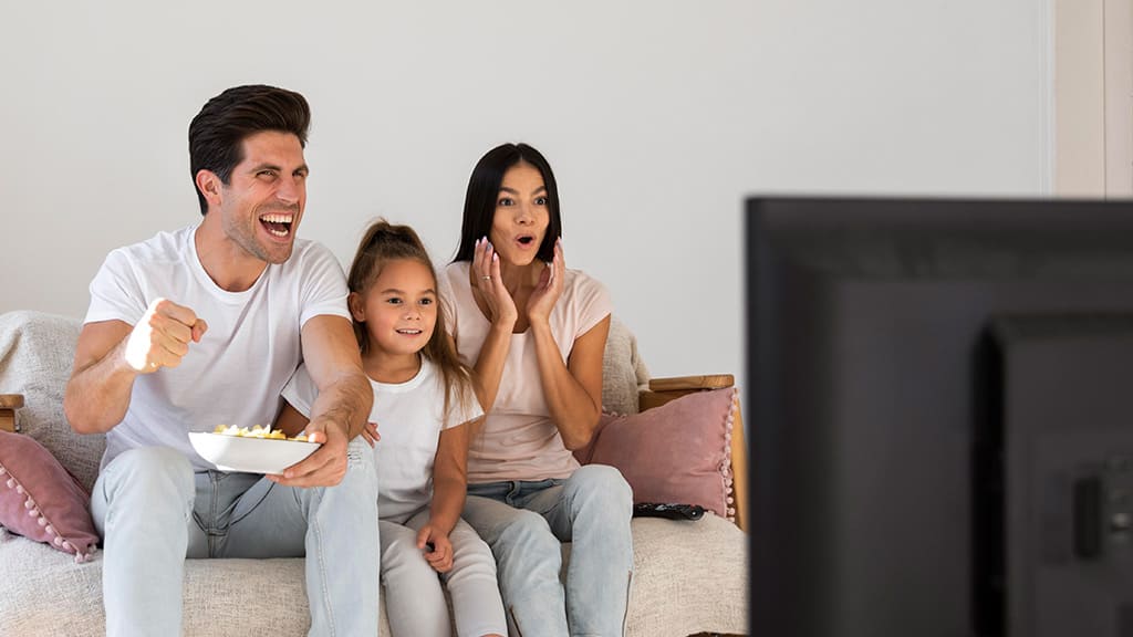 Uma família composta por um homem, uma mulher e uma criança, sentada no sofá e assistindo TV juntos.