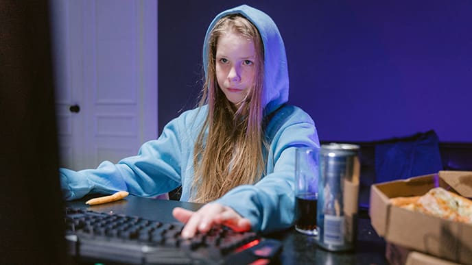 Uma gamer, de cabelos longos e blusa de frio, sentada em frente ao computador, jogando. Ao lado uma caixa de pizza e um refrigerante.