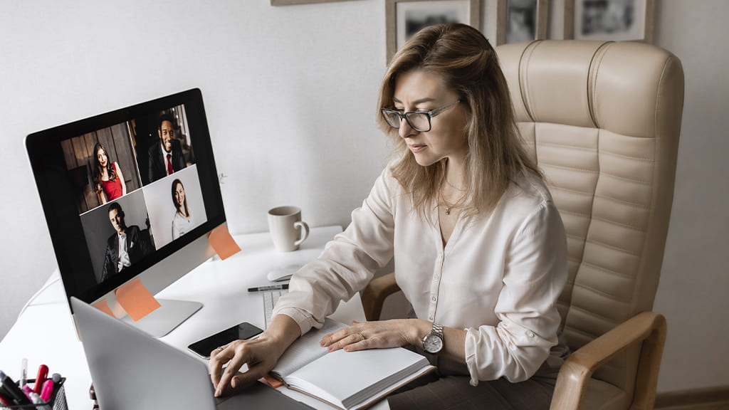 Uma mulher trabalhando em casa. Ela está sentada em uma mesa de escritório. A sua frente um notebook, uma tela de computador e uma agenda.