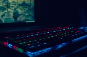 Em um ambiente escuro, um teclado luminoso, sendo que as teclas refletem luzes nas cores verde, amarelho, azul e vermelho. Ao fundo, a tela de um computador com a imagem de jogo.