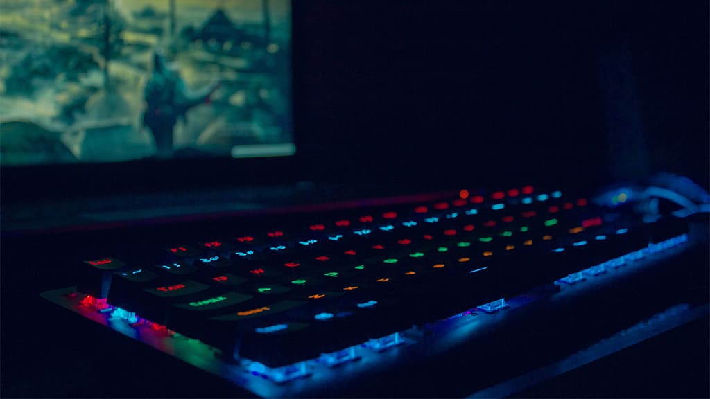 Em um ambiente escuro, um teclado luminoso, sendo que as teclas refletem luzes nas cores verde, amarelho, azul e vermelho. Ao fundo, a tela de um computador com a imagem de jogo.