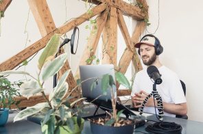 Um homem, sentado, gravando um podcast. A sala tem diversos equipamentos, uma estrutura de madeira e algumas plantas sobre a mesa.