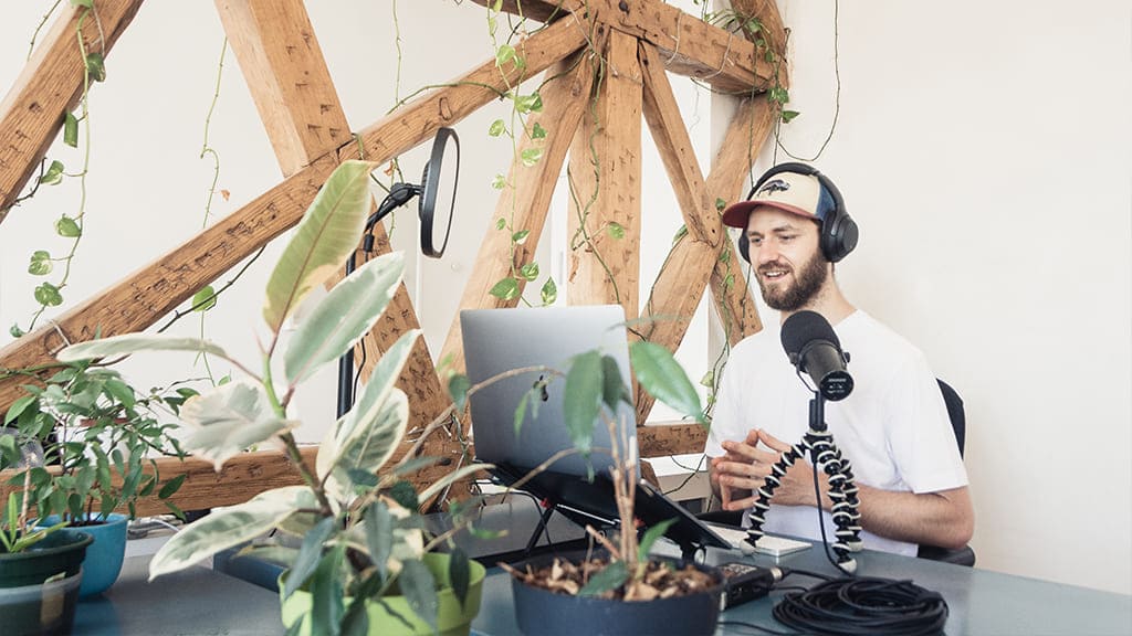 Um homem, sentado, gravando um podcast. A sala tem diversos equipamentos, uma estrutura de madeira e algumas plantas sobre a mesa.