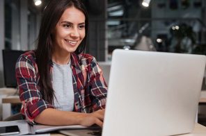 Uma mulher, no ambiente de trabalho, usando o notebook. Sobre a mesa, além da máquina, tem um celular, um lápisa e um caderna. Ela está com a semblante feliz por saber que está em um site seguro