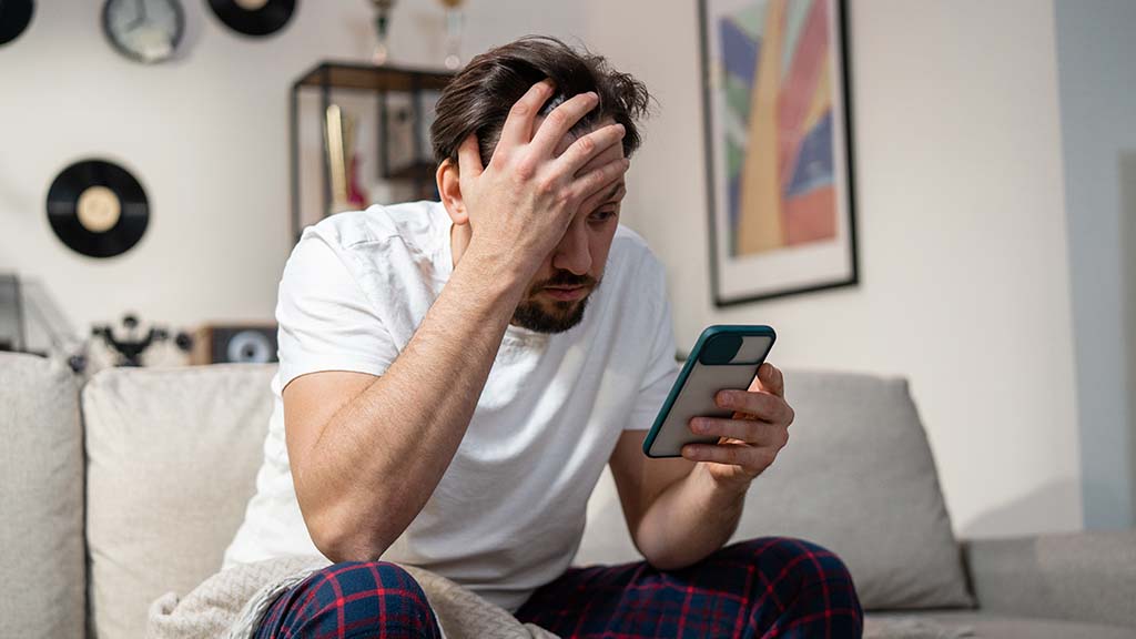 Um homem sentado no sofá. Uma mão está segurando o celular e a outra na cabeça. Ele está inconformado pois foi banido do WhatsApp