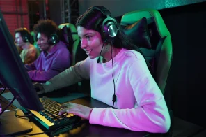 Uma menina gamer jogando no computador. Ela usa fone de ouvidos. Ao lado dela, outros gamers jogando.