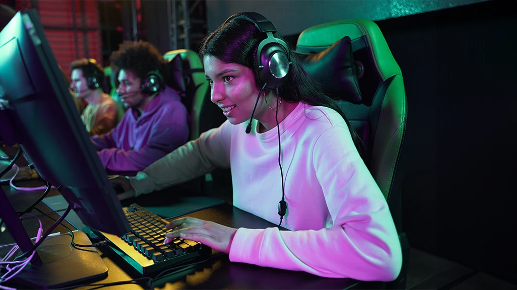 Uma menina gamer jogando no computador. Ela usa fone de ouvidos. Ao lado dela, outros gamers jogando.