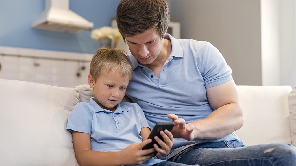 Pai e filho sentados no sofá, eles estão lendo no celular por meio do app do Skeelo