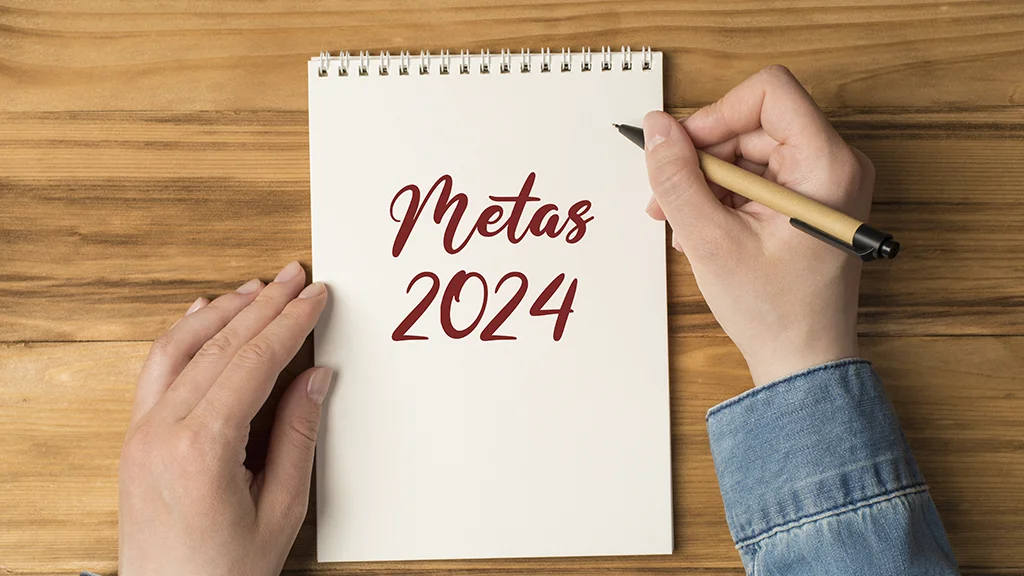 Uma pessoa com as mãos apoiadas sobre a mesa onde tem um bloco de notas. Ela segura uma lapiseira e escreveu "Metas 2024"