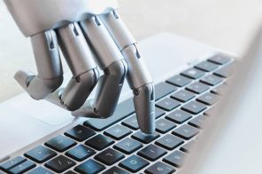 Na imagem, a mão de um robô digitando no teclado de um notebook.