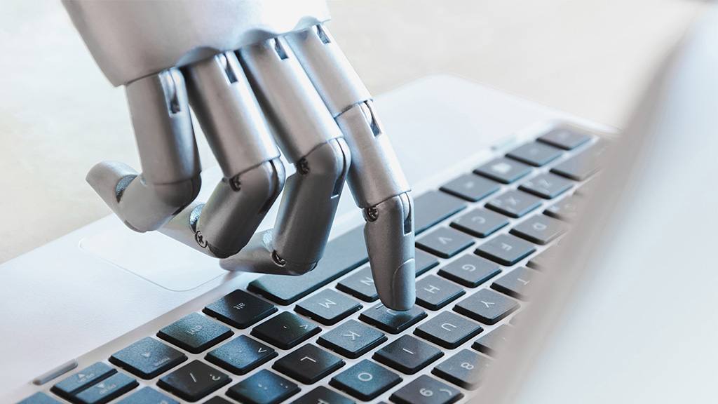 Na imagem, a mão de um robô digitando no teclado de um notebook.