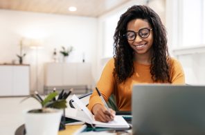 Na imagem uma adolescente, estudante. Ela tem um notebook e faz anotações, à caneta, em um caderno.