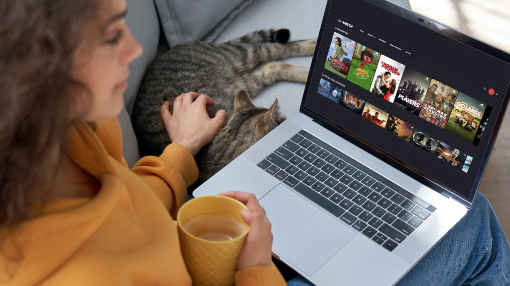 Imagem de uma mulher sentada no sofá. Ela tem um notebook no colo, com a tela da Watch Brasil. Com a mão direita ela segura uma caneca de leite e com a mão esquerda ela acaricia um gato.