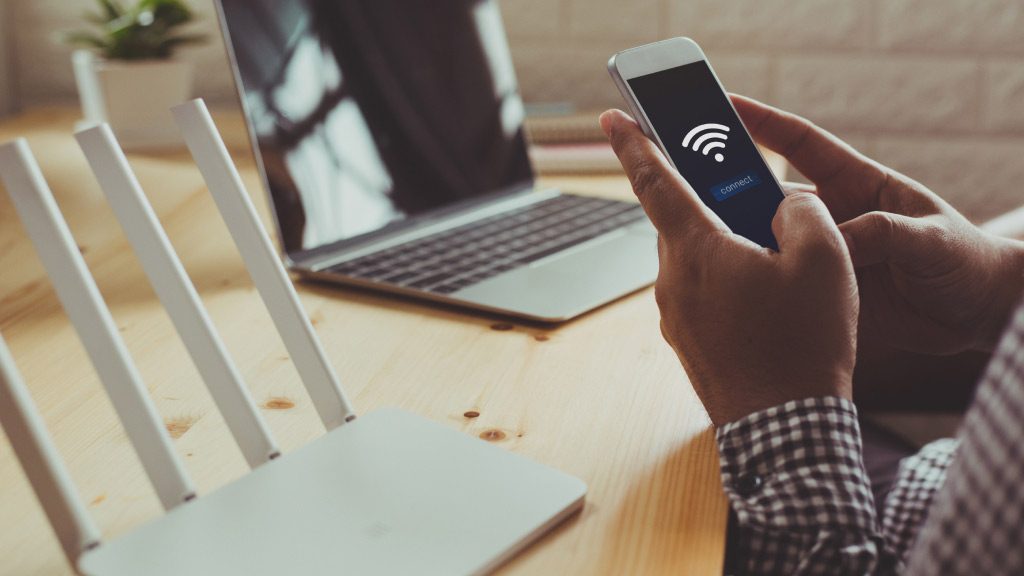 Na imagem, um homem próximo a mesa de escritório. Ele segura o celular e conecta o aparelho ao sinal do Wi-Fi. Sob a mesa, o roteador Wi-Fi e um notebook.