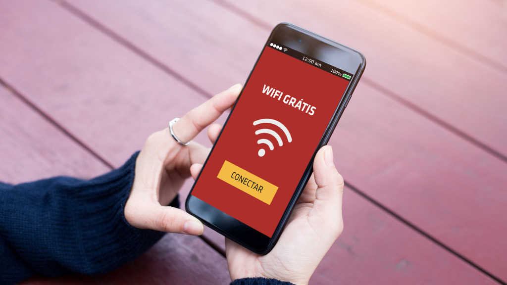 Na imagem, as mãos de uma pessoa segurando um celular. Na tela do aparelho, uma imagem com fundo vermelho e o seguinte texto: Wi-Fi grátis, conectar.