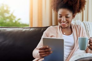Na imagem, uma mulher negra sentada no sofá. Ela segura uma caneca em uma das mãos e na outra um tablet.