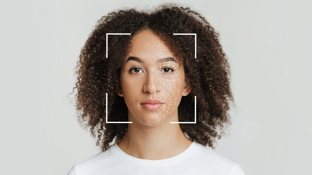 Na imagem, uma mulher negra, de cabelo solto até a altura dos ombros. Ela usa uma camiseta branca. O rosto dela está enquadrado em uma configuração de leitura de reconhecimento facial.