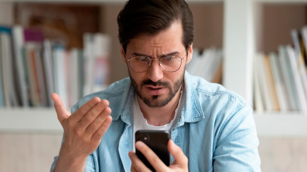Homem com camisa azul clara e camiseta branca, sentado com celular na mão, expressão de surpresa e uma mão levantada, preocupado por ter sido vítima de crime cibernético.