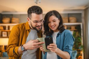 Casal de pé na sala conferindo livros sobre tecnologia no Skeelo. A moça segura o celular e sorri, vestindo blusa azul, enquanto o homem, de camisa mostarda e óculos redondos, observa a tela.
