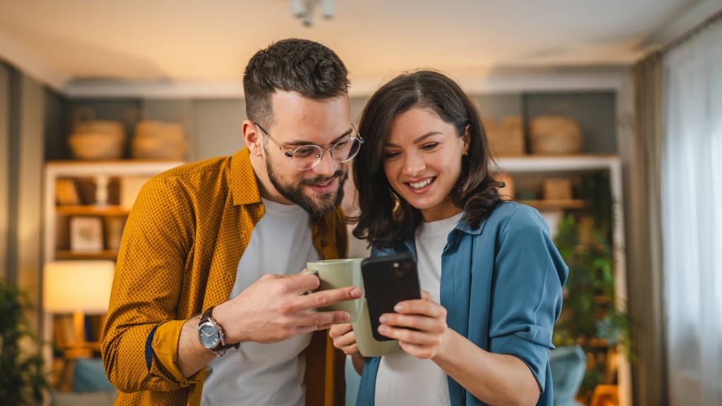 Casal de pé na sala conferindo livros sobre tecnologia no Skeelo. A moça segura o celular e sorri, vestindo blusa azul, enquanto o homem, de camisa mostarda e óculos redondos, observa a tela.