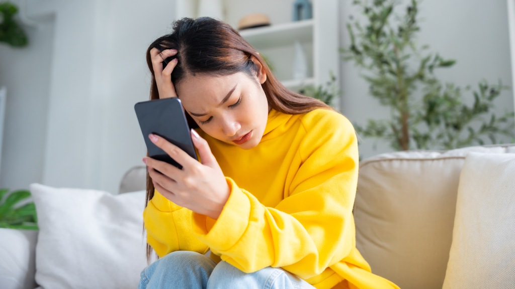 Uma moça vestindo um moletom amarelo. Ela está sentada no sofá e segura seu celular na mão esquerda. Ela está preocupada com o superaquecimento do aparelho.