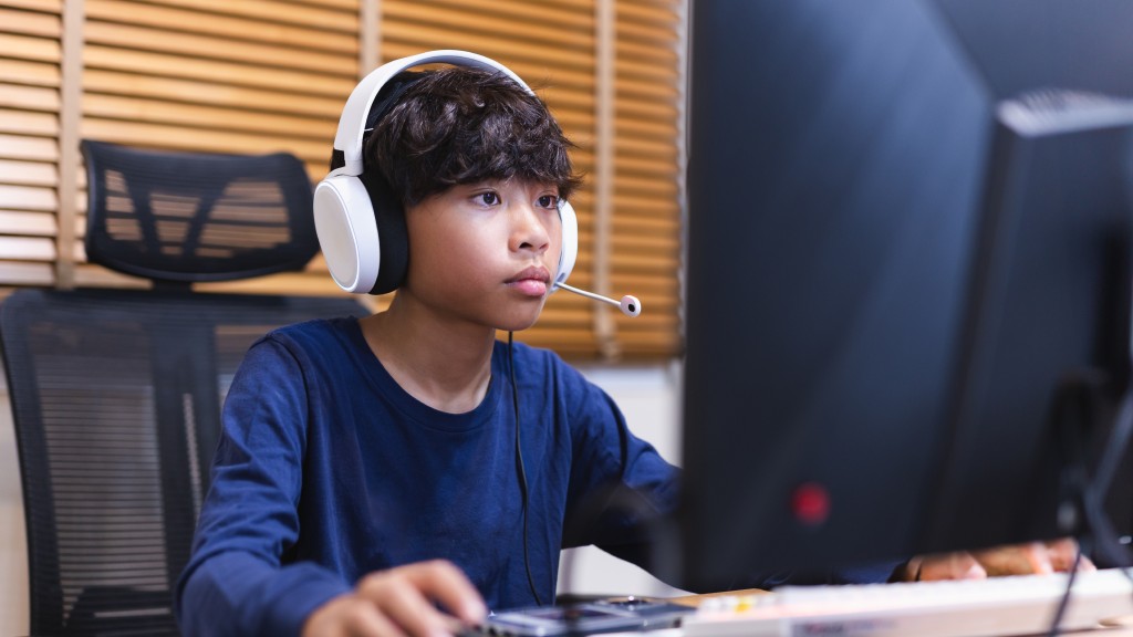 Menino gamer, vestido de azul e usando headphones brancos, sentados em frente ao computador jogando uma partida