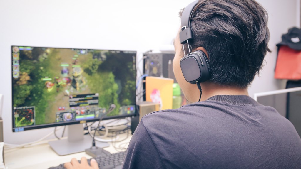 Menino gamer, vestindo camiseta azul marinho e usando headphones pretos, sentado em frente ao computador jogando uma partida