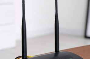 Roteador preto com luzes acesas sobre bancada de madeira, com parede branca e pequena prateleira com vaso de flor ao fundo, representando como reiniciar o roteador.