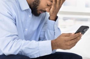 Homem de camisa azul clara e calça jeans, segurando o celular e apoiando a mão no rosto, demonstrando estresse com a internet oscilando.