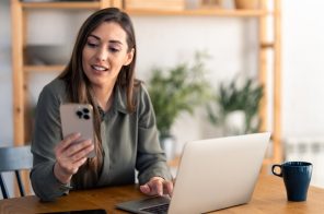 Mulher sentada à mesa com notebook e celular na mão, navegando com segurança graças à criptografia.