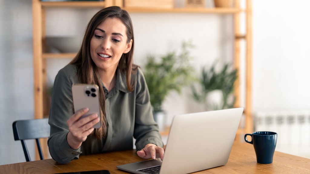 Mulher sentada à mesa com notebook e celular na mão, navegando com segurança graças à criptografia.