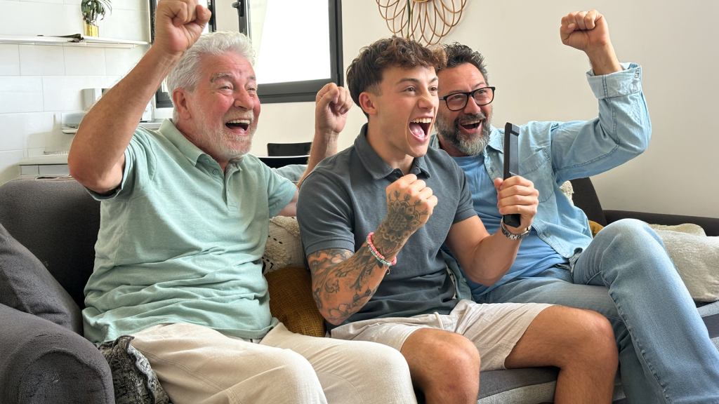 Três homens, um idoso, um de meia idade e um jovem, vibrando juntos, sentados no sofá, ao assistir uma partida da NBA com o app NBA Team Pass.