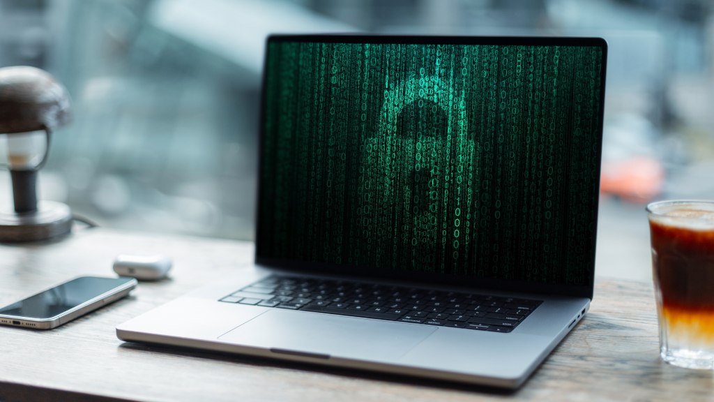 Notebook sobre a mesa ao lado de celular, fones e copo, com cadeado na tela simbolizando dados criptografados.