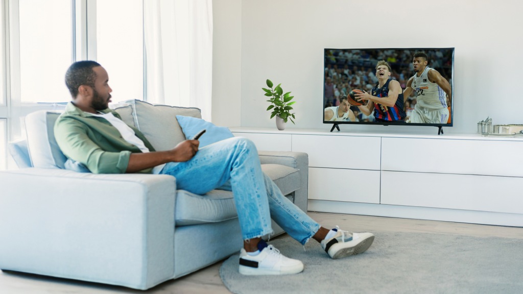 Homem jovem negro sentado em um sofá cinza, usando camisa verde e calças jeans, assistindo a uma partida da NBA