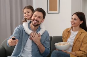 Família feliz com marido, esposa e filha apoiada nas costas do pai; mãe segura um balde de pipoca e pai um controle remoto enquanto assistem ao Paramount+.