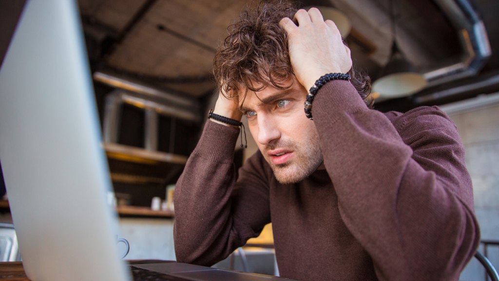 Homem jovem de blusa marrom sentado à frente do notebook com as mãos na cabeça, preocupado por ter seus dados vazados na internet.