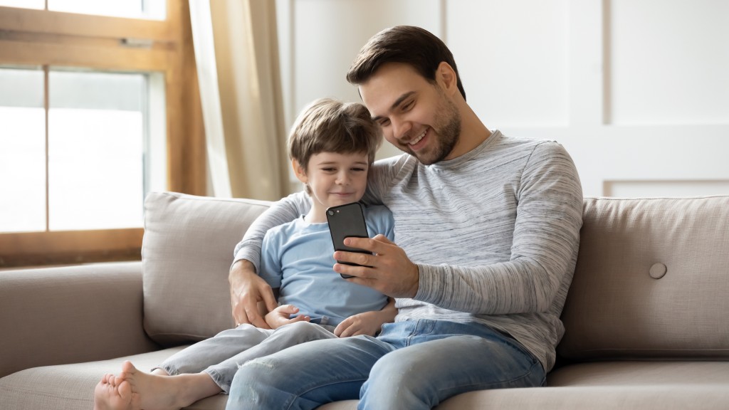Homem de camisa cinza e filho de camiseta azul sentados no sofá, sorrindo enquanto acessam aplicativo para crianças no celular.