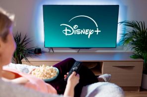 Mulher sentada no sofá com pipoca no colo e controle remoto na mão assistindo Disney+ em sua TV.