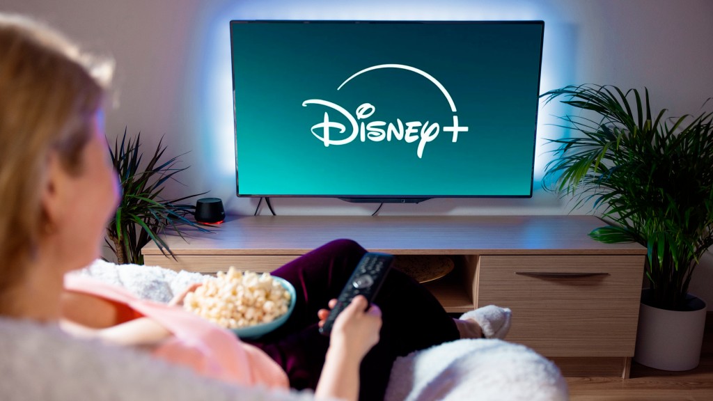 Mulher sentada no sofá com pipoca no colo e controle remoto na mão assistindo Disney+ em sua TV.