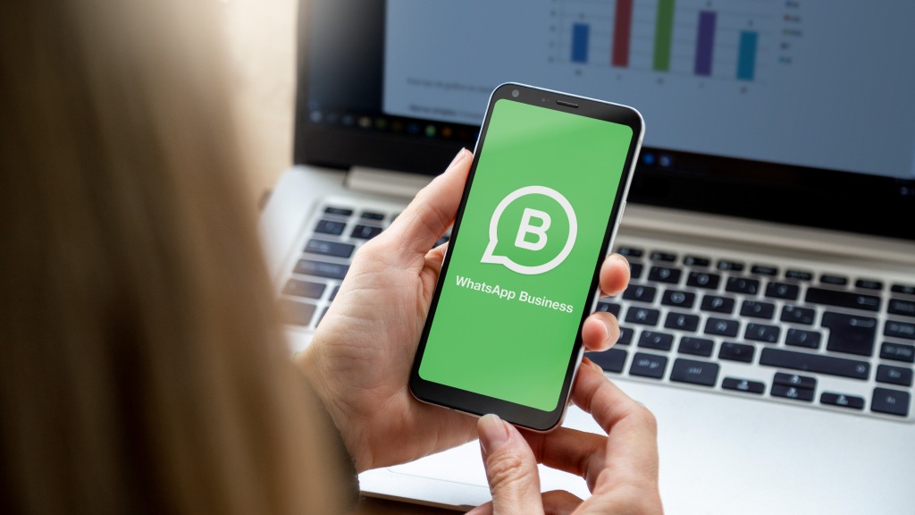 Pessoa segurando um celular com as duas mãos com a tabela exibindo o logo do WhatsApp Business. Ao fundo da imagem, há um computador com gráfico em barras na tela.