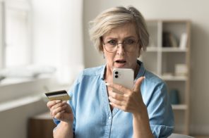 Mulher de meia-idade com cabelos curtos grisalhos e camisa azul, espantada com dados vazados na internet enquanto segura iPhone branco e cartão de crédito.