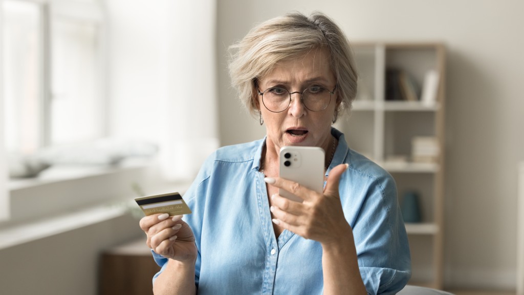 Mulher de meia-idade com cabelos curtos grisalhos e camisa azul, espantada com dados vazados na internet enquanto segura iPhone branco e cartão de crédito.